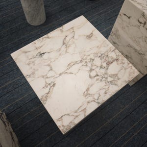May include: A white marble square table top with gray veining. The table top is sitting on a gray carpet.