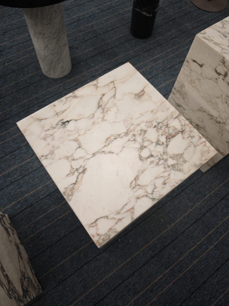 May include: A white marble square table top with gray veining. The table top is sitting on a gray carpet.