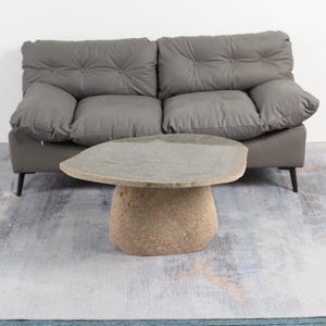 Puede incluir: Una mesa de café de piedra rústica con una parte superior ovalada irregular y una base redondeada. La mesa está hecha de piedra natural de color marrón claro y gris. Está sentada sobre una alfombra gris y azul frente a un sofá gris.