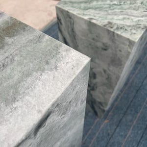Close-up top view of two green marble pedestal stands showing polished surfaces and cloudy veining patterns. The subtle blend of green, gray, and white tones lends each piece a soft, natural feel. These versatile plinths can function as plant stands, decorative side tables, or sculptural display columns. Each pedestal is hand-finished to highlight the beauty of the stone, making them a luxurious and functional furniture option for interior styling.