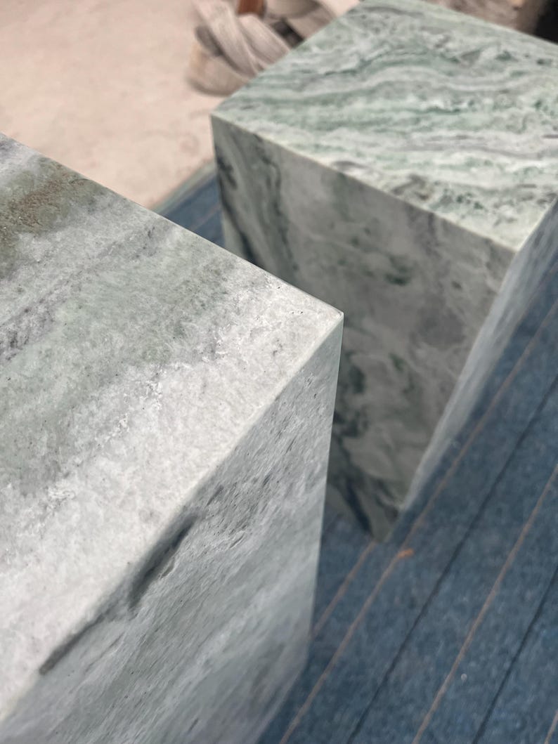 Close-up top view of two green marble pedestal stands showing polished surfaces and cloudy veining patterns. The subtle blend of green, gray, and white tones lends each piece a soft, natural feel. These versatile plinths can function as plant stands, decorative side tables, or sculptural display columns. Each pedestal is hand-finished to highlight the beauty of the stone, making them a luxurious and functional furniture option for interior styling.
