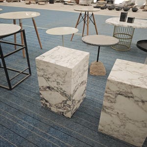 May include: Two white marble square pedestals with gray veining, one on top of the other, on a blue and white striped floor.