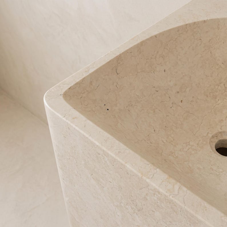 May include: A close-up of a beige stone sink with a rounded corner. The sink has a rectangular basin with a drain hole. The stone has a natural, veined pattern. The background is a matching beige wall.