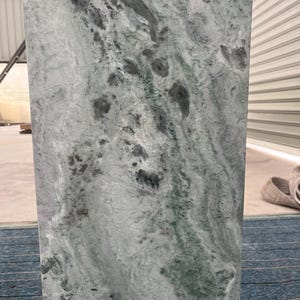 Sunlit green marble pedestal stand with prominent veining in light green and white. Placed on a blue carpet near a white wall, the stone’s natural texture is emphasized by direct sunlight. The pedestal’s clean shape and solid construction make it a beautiful base for art or floral installations. Whether styled in a gallery, studio, or refined home, this pedestal adds architectural charm and luxury through its organic material and contemporary form.