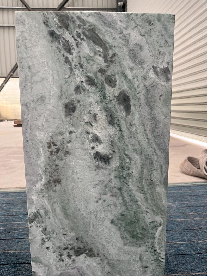 Sunlit green marble pedestal stand with prominent veining in light green and white. Placed on a blue carpet near a white wall, the stone’s natural texture is emphasized by direct sunlight. The pedestal’s clean shape and solid construction make it a beautiful base for art or floral installations. Whether styled in a gallery, studio, or refined home, this pedestal adds architectural charm and luxury through its organic material and contemporary form.
