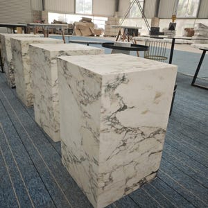 May include: Four white marble cube-shaped stools with gray veining. The stools are arranged in a row on a blue and gray striped carpet.