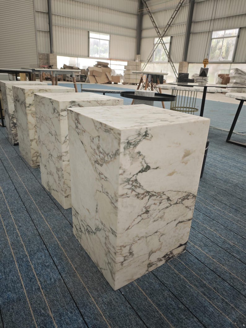 May include: Four white marble cube-shaped stools with gray veining. The stools are arranged in a row on a blue and gray striped carpet.