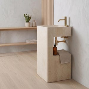 May include: A modern, beige stone pedestal sink with a gold faucet and plumbing. A small shelf holds a brown bottle and a folded towel. The sink is in a bathroom setting with a wooden shelf and a plant.