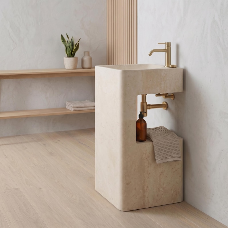 May include: A modern, beige stone pedestal sink with a gold faucet and plumbing. A small shelf holds a brown bottle and a folded towel. The sink is in a bathroom setting with a wooden shelf and a plant.