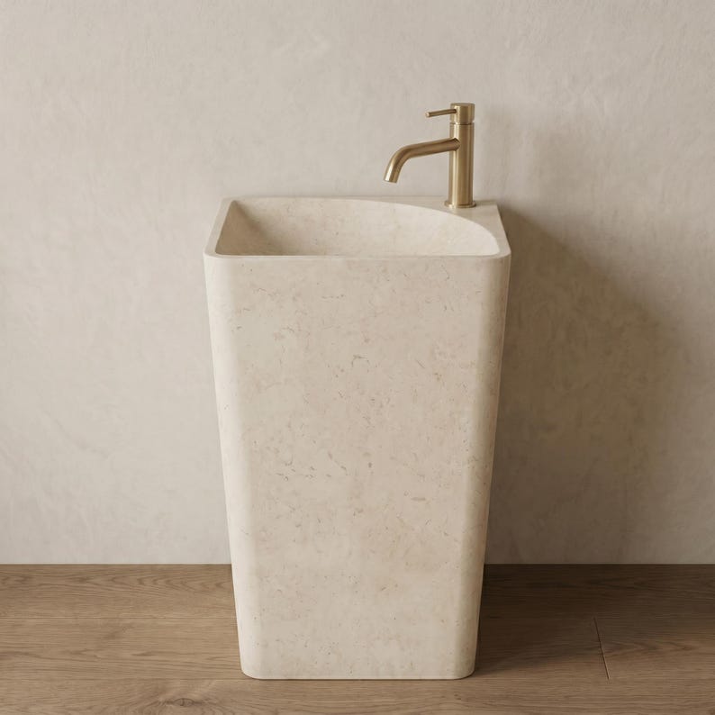 May include: A freestanding, rectangular beige stone sink with a curved top edge. A gold-colored faucet is mounted on the sink's edge. The sink is set against a neutral-toned wall and sits on a wooden floor. The sink has a modern, minimalist design.