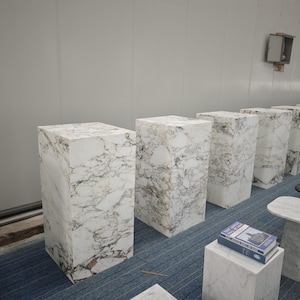 May include: A row of white marble pedestals of varying heights, arranged in a room with a white wall and a blue carpet. The pedestals are made of white marble with gray veining.