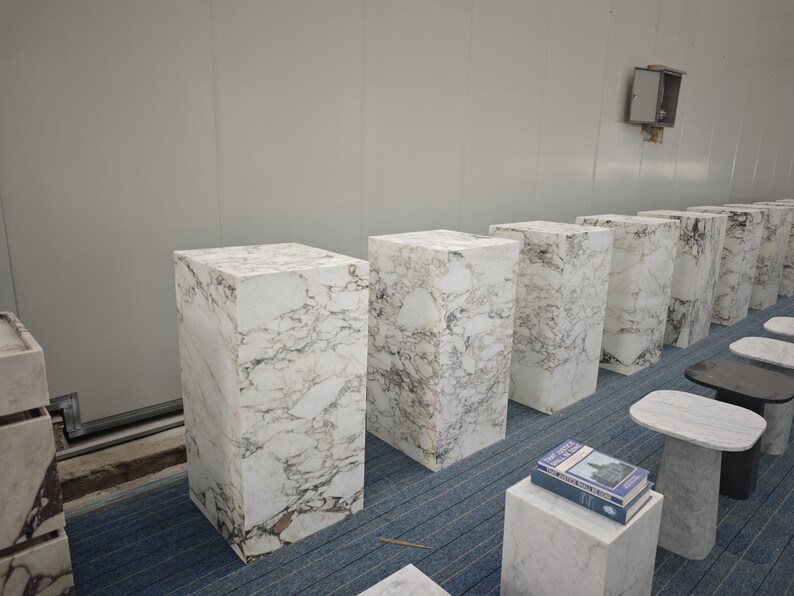 May include: A row of white marble pedestals of varying heights, arranged in a room with a white wall and a blue carpet. The pedestals are made of white marble with gray veining.