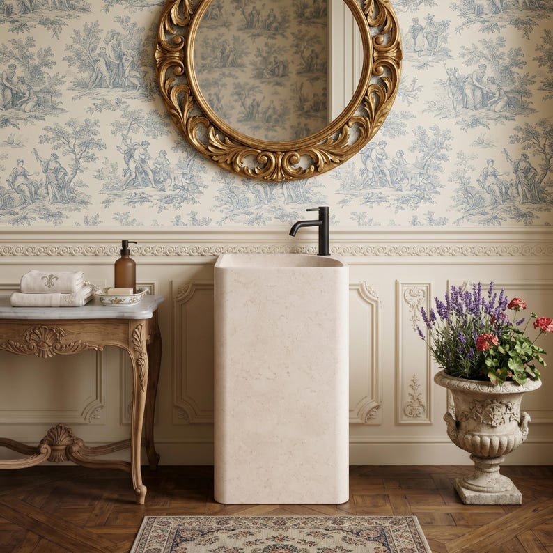May include: A bathroom scene featuring a beige pedestal sink with a black faucet, a gold-framed mirror, and a vintage-style table with towels and a soap dish. The wallpaper has a blue and white pastoral design, and a decorative urn holds flowers.