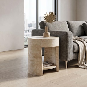 May include: A beige, cylindrical side table with a drawer and a lower shelf. A small, beige vase with a dried palm leaf sits on top. The table is next to a gray sofa. A book titled "Scandinavian Design" is on the shelf.