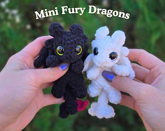 May include: Two small crocheted plush toys labeled Mini Fury Dragons. One is black with large yellow eyes and a small red tail accent, and the other is white with large black eyes. Both have rounded ears, small wings, and textured yarn bodies.