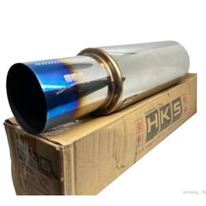 May include: A shiny chrome exhaust silencer with a blue-hued outlet. The silencer is cylindrical with a gold-coloured band near the tip. The box below the silencer has the HKS logo printed on it.