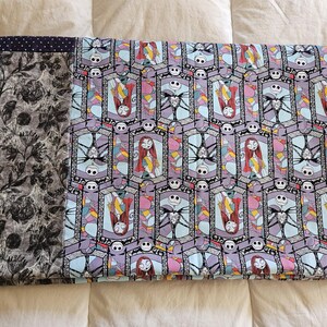 May include: A blue and purple fabric blanket with a repeating pattern of stained glass windows featuring Jack Skellington and Sally from The Nightmare Before Christmas. The blanket has a black and white floral border.