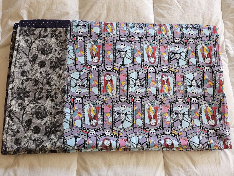 May include: A blue and purple fabric blanket with a repeating pattern of stained glass windows featuring Jack Skellington and Sally from The Nightmare Before Christmas. The blanket has a black and white floral border.