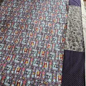 May include: A quilt with a patchwork design featuring a repeating pattern of Jack Skellington and Sally from The Nightmare Before Christmas. The quilt is made of different fabrics, including a purple fabric with a white polka dot pattern.
