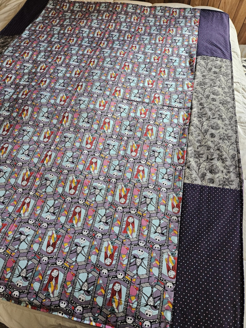 May include: A quilt with a patchwork design featuring a repeating pattern of Jack Skellington and Sally from The Nightmare Before Christmas. The quilt is made of different fabrics, including a purple fabric with a white polka dot pattern.