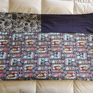 May include: A purple and blue quilt with a repeating pattern of Jack Skellington and Sally from the Nightmare Before Christmas. The quilt is made of three different fabrics, one with a black and white floral pattern, one with a solid purple fabric with white polka dots, and one with the repeating pattern of Jack and Sally.