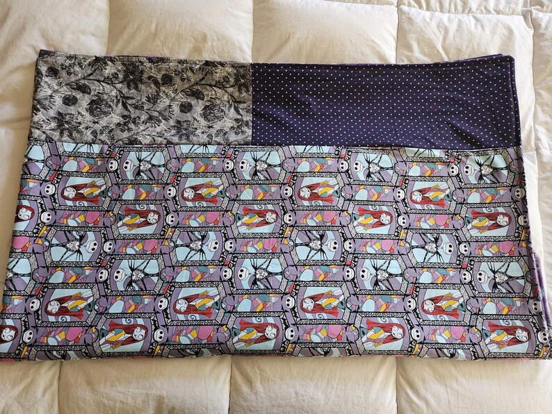 May include: A purple and blue quilt with a repeating pattern of Jack Skellington and Sally from the Nightmare Before Christmas. The quilt is made of three different fabrics, one with a black and white floral pattern, one with a solid purple fabric with white polka dots, and one with the repeating pattern of Jack and Sally.