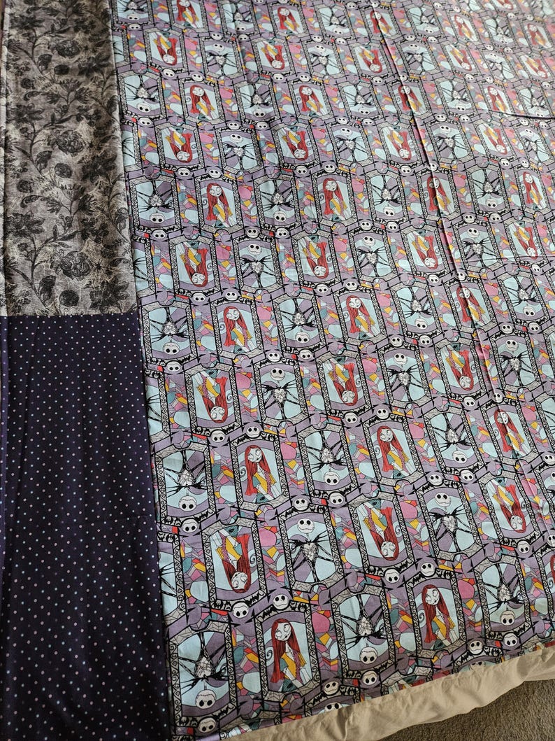 May include: A quilt with a patchwork design featuring a black and white floral pattern, a black fabric with white polka dots, and a colorful fabric with a repeating pattern of Jack Skellington and Sally from The Nightmare Before Christmas.