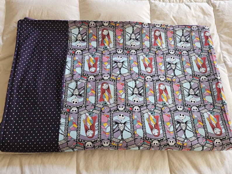 May include: A purple and blue patterned blanket with a black and white polka dot backing. The pattern features a repeating design of Jack Skellington and Sally from the Nightmare Before Christmas.