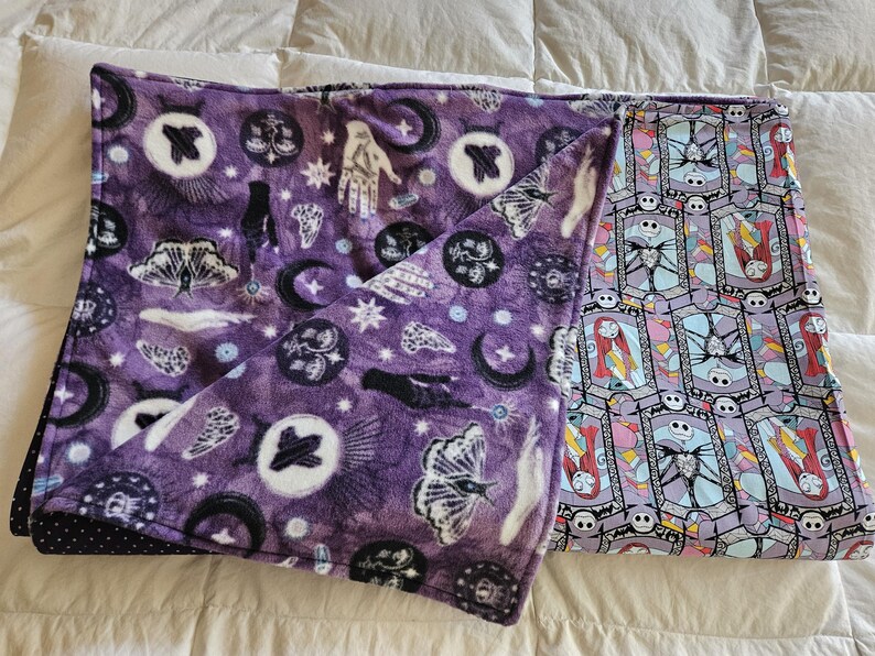 May include: A purple fleece blanket with a black and white pattern of moons, stars, hands, and butterflies. The other side of the blanket is a light purple and blue pattern with a repeated image of a character from the movie The Nightmare Before Christmas.