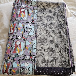 May include: A folded purple and gray fleece blanket with a stained glass pattern featuring Jack Skellington and Sally from The Nightmare Before Christmas. The blanket has a black and white floral pattern on one side and a black and white polka dot pattern on the other side.