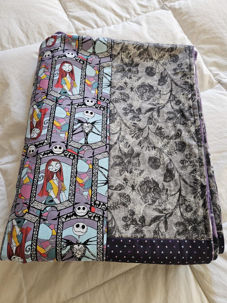 May include: A folded purple and gray fleece blanket with a stained glass pattern featuring Jack Skellington and Sally from The Nightmare Before Christmas. The blanket has a black and white floral pattern on one side and a black and white polka dot pattern on the other side.