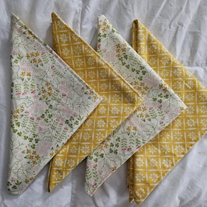 May include: Four folded cloth napkins. Two napkins feature a yellow floral pattern, while the other two have a white background with pink and green floral and leaf designs. The napkins are arranged on a white surface.