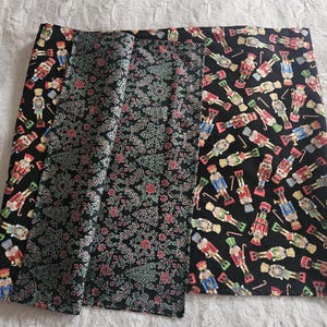 May include: A folded fabric with three distinct patterns. One section features a black background with colorful nutcracker figures. Another has a black background with a floral design in pink and green. The third section has a black background with a floral design.