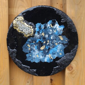 May include: A round resin art piece with a black background and a blue and white abstract design with gold accents.