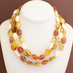 May include: A three-strand necklace displayed on a white bust. The necklace features oval-shaped beads in shades of yellow, cream, brown, and amber. The beads are strung together, creating a layered effect. The background is a solid brown.
