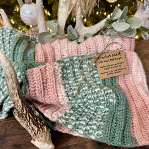 May include: A handmade crochet scarf in shades of pink, green, and white. The scarf is made of a wool and acrylic blend and features a ribbed design. A tag reads "Crochet Scarf". The scarf is displayed with decorative antlers and greenery.