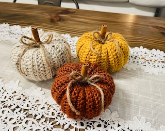 May include: Three handmade crochet pumpkins in cream, mustard yellow, and rust orange. Each pumpkin has a cinnamon stick stem and is tied with twine. The pumpkins are arranged on a white lace runner, perfect for fall decor.
