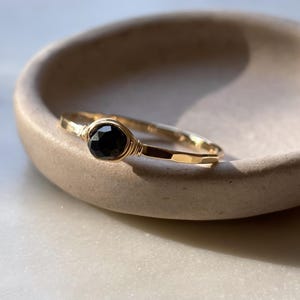 May include: A gold ring featuring a single, round, faceted black gemstone set in a wire-wrapped bezel. The band has a hammered, textured finish, creating a reflective surface that catches the light. The design is simple and focused on the contrast between the dark stone and the metallic gold band.
