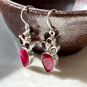May include: A pair of silver earrings with a teardrop-shaped, deep pink gemstone at the bottom and three smaller, round, dark red gemstones above. The earrings are set against a blurred background.