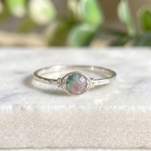Puede incluir: Anillo de plata con una gema facetada de forma ovalada. La gema presenta tonos verdes y rosas, engarzada en un diseño de alambre. La banda tiene una textura martillada, añadiendo un toque rústico a la joya.
