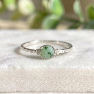 Könnte beinhalten: Ein silberner Ring mit einem hellgrünen, facettierten Edelstein in der Mitte. Der Edelstein wird von einer Drahtwicklung gehalten. Das Band hat eine gehämmerten Textur. Der Ring liegt auf einer weißen Oberfläche.
