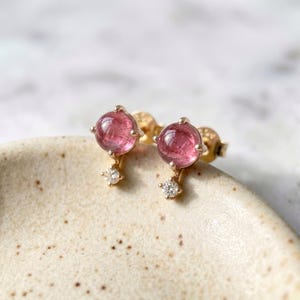 Può includere: Un paio di orecchini a lobo in oro con cabochon di pietre preziose rosa e piccoli accenti di diamanti. Gli orecchini hanno una chiusura a perno e un design delicato ed elegante. Le pietre preziose sono di un rosa vibrante.