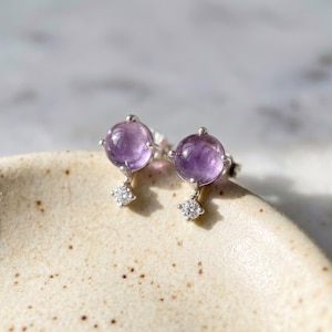 Puede incluir: Un par de pendientes de botón con piedras redondas de amatista de color púrpura claro. Cada pendiente tiene un pequeño acento colgante en forma de diamante. Los pendientes están engarzados en un metal plateado.