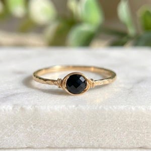 May include: A gold ring featuring a hammered band and a round, faceted black gemstone. The gemstone is secured with wire wrapping. The ring is set against a white surface with blurred green foliage in the background.