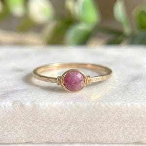 May include: A gold-colored ring with a hammered band and a round, faceted, pink gemstone. The gemstone is secured with gold wire. The ring is set against a white background with blurred green foliage.