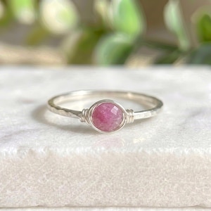 May include: A delicate silver ring with a faceted, round, pink gemstone at the center. The gemstone is held in place by wire wrapping. The ring band has a hammered texture, adding a rustic touch to the jewelry.