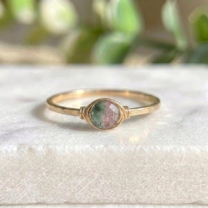 May include: A delicate gold ring with a round, multi-colored gemstone. The gemstone features green and pink hues, set in a wire-wrapped design. The ring's band has a hammered texture, adding a unique touch to this piece of jewelry.