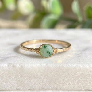 May include: A gold ring featuring a light green, faceted gemstone. The stone is secured with a wire wrap. The ring's band has a hammered texture. The ring is displayed on a white surface. The ring's diameter is approximately 1.7 cm.