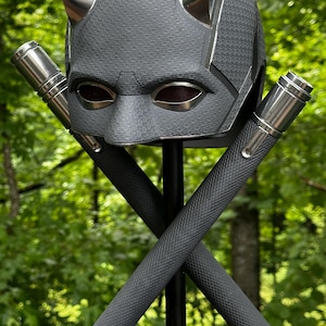 May include: A dark gray helmet with silver accents and curved horns, resembling a superhero's mask. Two gray batons with red tips are crossed in front of the helmet, creating a striking visual.