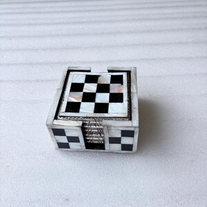 May include: A set of square coasters with a black and white checkerboard pattern on top. The coasters are housed in a matching box with a similar design. The box and coasters have a mother-of-pearl inlay.